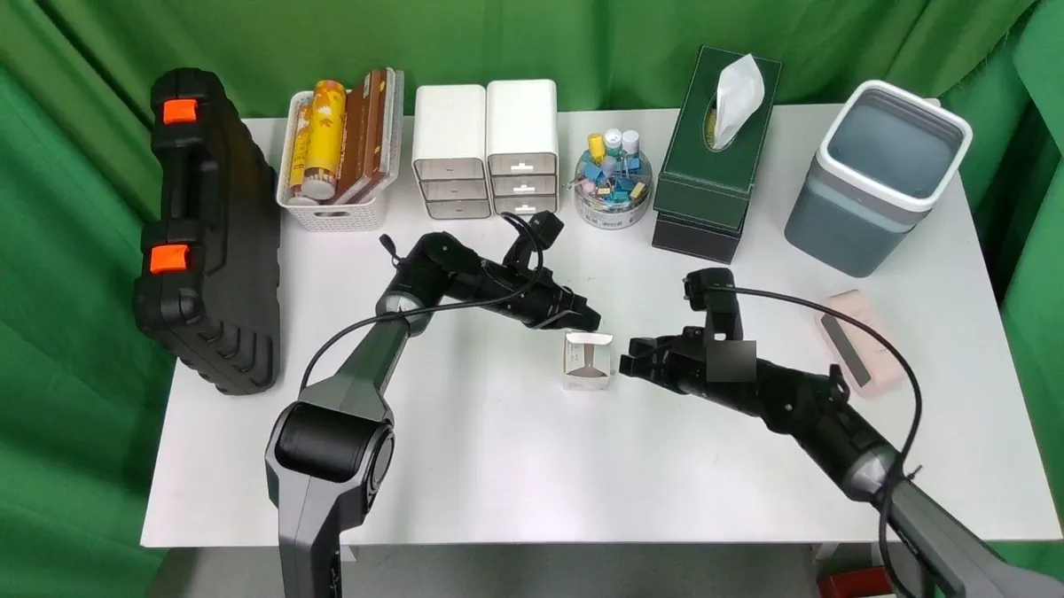 Top camera view of two robot arms with parallel grippers handling a small box on a desk